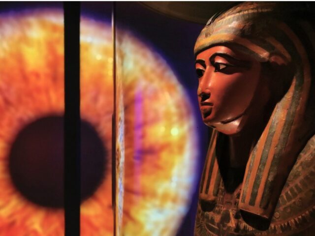 Ett stort öga med eldfärgad iris i bakgrunden. Till höger i bild överdelen på en antik egyptisk kista.